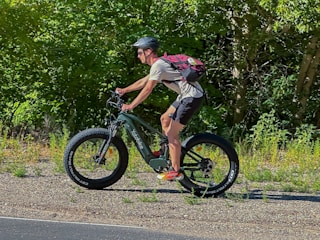 A person is riding a green mountain bike on a gravel path with large, thick tires. The individual is wearing a helmet, a red backpack, and athletic clothing. The background is filled with dense green foliage, suggesting a lush and natural environment.