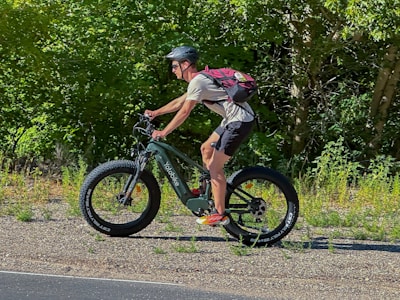 A person is riding a green mountain bike on a gravel path with large, thick tires. The individual is wearing a helmet, a red backpack, and athletic clothing. The background is filled with dense green foliage, suggesting a lush and natural environment.
