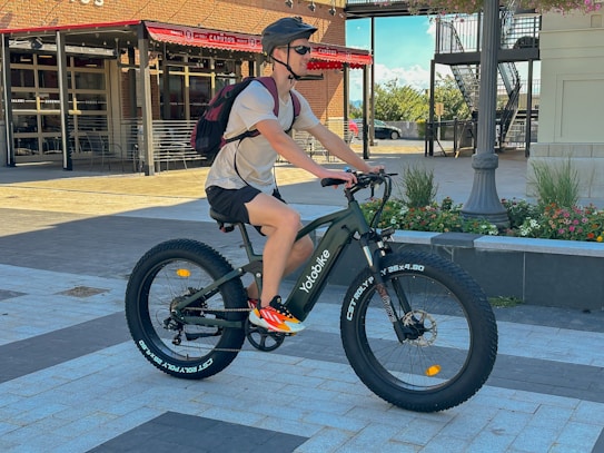 A person wearing a helmet and sunglasses rides a fat-tire bike on a paved area near a red-bricked building with a restaurant. They carry a backpack and wear casual clothing. The background includes a lamp post and flowerbeds.