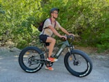 A person is riding an electric bike on a forest trail, surrounded by lush green trees. They are wearing a helmet, sunglasses, a t-shirt, shorts, and colorful sneakers. A backpack is slung over their shoulders.