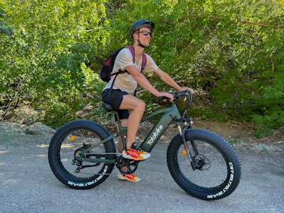 A person is riding an electric bike on a forest trail, surrounded by lush green trees. They are wearing a helmet, sunglasses, a t-shirt, shorts, and colorful sneakers. A backpack is slung over their shoulders.