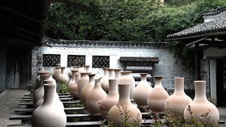 A collection of large, traditional clay vases are arranged in rows on wooden planks outside a rustic building. The vases are unglazed and have a natural, earthy tone. In the background, there are whitewashed walls with lattice windows and lush green foliage. The setting appears tranquil and steeped in cultural heritage.
