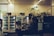 A dimly lit indoor space with two refrigerated display units containing various packaged goods. A large leafy plant decorates the area between the refrigerators. In the foreground, a counter with kitchen appliances and bottles is visible. The setting appears to be a self-service or small convenience area with a calm ambiance.