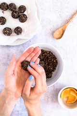 A pair of hands is forming a small, round, dark energy ball from a mixture in a bowl. Several completed energy balls rest on a piece of parchment paper on a plate. A small spoon lies nearby on the textured surface, and a cup with orange slices is visible in the bottom corner.