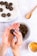 A pair of hands is forming a small, round, dark energy ball from a mixture in a bowl. Several completed energy balls rest on a piece of parchment paper on a plate. A small spoon lies nearby on the textured surface, and a cup with orange slices is visible in the bottom corner.