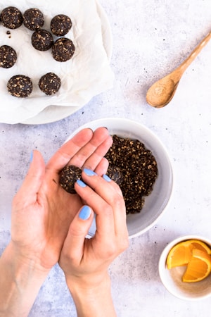 A pair of hands is forming a small, round, dark energy ball from a mixture in a bowl. Several completed energy balls rest on a piece of parchment paper on a plate. A small spoon lies nearby on the textured surface, and a cup with orange slices is visible in the bottom corner.