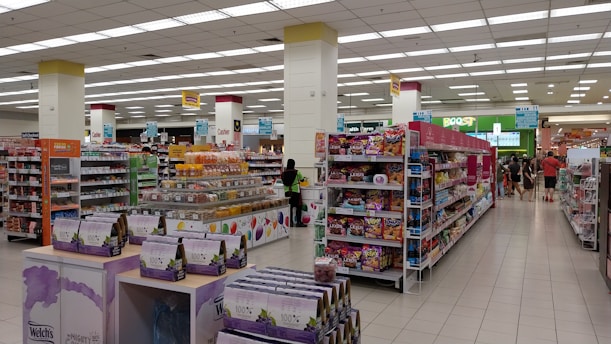 A spacious, well-lit supermarket aisle features neatly arranged shelves stocked with various food products, including snacks, beverages, and packaged goods. The store's layout is organized, with clear signage indicating different sections and a visible checkout area in the distance. Customers can be seen browsing and shopping.