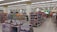 A spacious, well-lit supermarket aisle features neatly arranged shelves stocked with various food products, including snacks, beverages, and packaged goods. The store's layout is organized, with clear signage indicating different sections and a visible checkout area in the distance. Customers can be seen browsing and shopping.