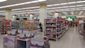 A spacious, well-lit supermarket aisle features neatly arranged shelves stocked with various food products, including snacks, beverages, and packaged goods. The store's layout is organized, with clear signage indicating different sections and a visible checkout area in the distance. Customers can be seen browsing and shopping.