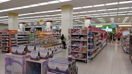 A spacious, well-lit supermarket aisle features neatly arranged shelves stocked with various food products, including snacks, beverages, and packaged goods. The store's layout is organized, with clear signage indicating different sections and a visible checkout area in the distance. Customers can be seen browsing and shopping.