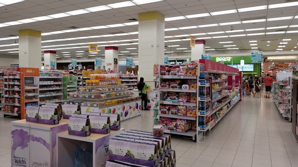 A spacious, well-lit supermarket aisle features neatly arranged shelves stocked with various food products, including snacks, beverages, and packaged goods. The store's layout is organized, with clear signage indicating different sections and a visible checkout area in the distance. Customers can be seen browsing and shopping.