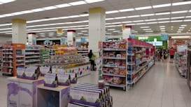 A spacious, well-lit supermarket aisle features neatly arranged shelves stocked with various food products, including snacks, beverages, and packaged goods. The store's layout is organized, with clear signage indicating different sections and a visible checkout area in the distance. Customers can be seen browsing and shopping.