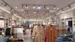 A clothing store interior featuring multiple racks with various garments. The ceiling has track lighting, and the walls are lined with shelves displaying folded clothes. Brick accents give an industrial feel, and the aisles are organized with apparel such as dresses, shirts, and jeans.