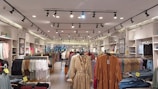A clothing store interior featuring multiple racks with various garments. The ceiling has track lighting, and the walls are lined with shelves displaying folded clothes. Brick accents give an industrial feel, and the aisles are organized with apparel such as dresses, shirts, and jeans.