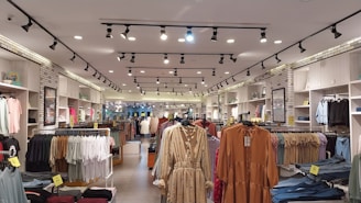 A clothing store interior featuring multiple racks with various garments. The ceiling has track lighting, and the walls are lined with shelves displaying folded clothes. Brick accents give an industrial feel, and the aisles are organized with apparel such as dresses, shirts, and jeans.