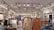 A clothing store interior featuring multiple racks with various garments. The ceiling has track lighting, and the walls are lined with shelves displaying folded clothes. Brick accents give an industrial feel, and the aisles are organized with apparel such as dresses, shirts, and jeans.