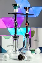 Two hookahs are prominently displayed, each with a sleek transparent base and a colorful neck piece. They are set against a modern backdrop with multicolored LED triangle lights. Several white spheres and boxes labeled 'Moze Sphere 2' are arranged around the hookahs, enhancing the contemporary and stylish look.