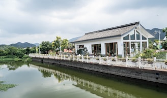 A modern house with large windows and a traditional roof design is situated by a calm river. The property is bordered by a white fence and there is lush greenery around. The sky is overcast, and the surrounding landscape includes rolling hills.
