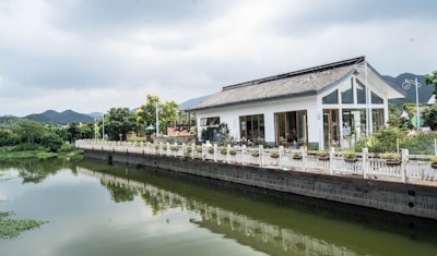 A modern house with large windows and a traditional roof design is situated by a calm river. The property is bordered by a white fence and there is lush greenery around. The sky is overcast, and the surrounding landscape includes rolling hills.
