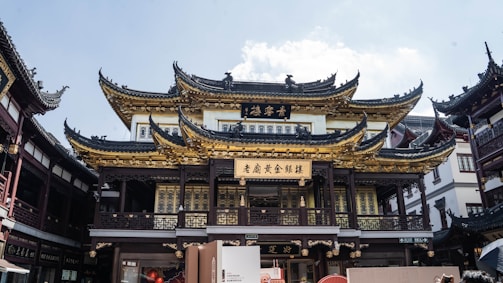 Intricately designed traditional Chinese architecture with ornate, curved rooflines. The building is adorned with gold and dark wood elements, showcasing detailed craftsmanship. Surrounding structures complement the central building’s style, creating a cohesive aesthetic in an urban setting.