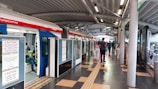 A modern train station platform with a train on the left side. Several passengers are seen either boarding the train or walking on the platform. There are caution signs on the train doors indicating they are under construction. The platform has a tiled floor and a curved metal roof. Lighting is provided by numerous ceiling fixtures.