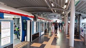 A modern train station platform with a train on the left side. Several passengers are seen either boarding the train or walking on the platform. There are caution signs on the train doors indicating they are under construction. The platform has a tiled floor and a curved metal roof. Lighting is provided by numerous ceiling fixtures.