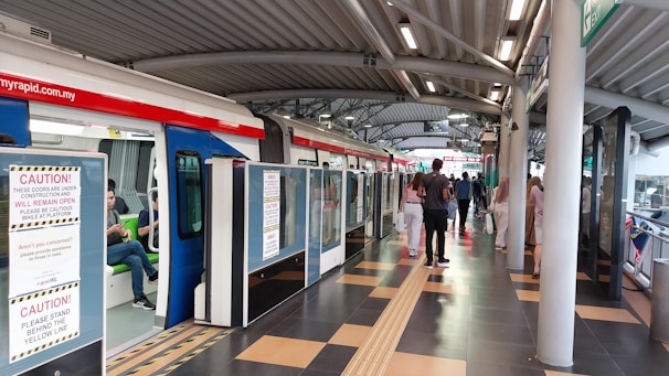 A modern train station platform with a train on the left side. Several passengers are seen either boarding the train or walking on the platform. There are caution signs on the train doors indicating they are under construction. The platform has a tiled floor and a curved metal roof. Lighting is provided by numerous ceiling fixtures.