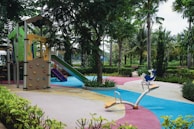 A colorful playground with a climbing structure, slide, and seesaw is surrounded by trees and greenery. The ground is covered with vibrant, interweaving paths of blue, pink, yellow, and gray. Benches are situated along the pathways encircled by bushes and palms that contribute to a lush, inviting environment.