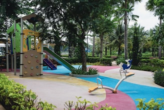 A colorful playground with a climbing structure, slide, and seesaw is surrounded by trees and greenery. The ground is covered with vibrant, interweaving paths of blue, pink, yellow, and gray. Benches are situated along the pathways encircled by bushes and palms that contribute to a lush, inviting environment.