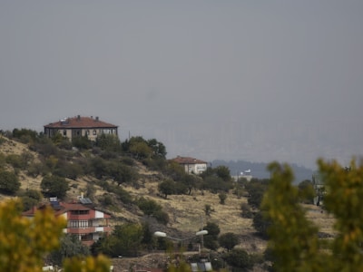 A rural landscape features houses with red roofs situated on a hilly terrain with sparse trees and bushes. The background shows a hazy city skyline, giving a contrast between nature and urban development.