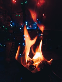 Flames rise from a small open fire in a dark setting, surrounded by multi-colored fairy lights offering a festive atmosphere. A bottle with partially visible text stands nearby.