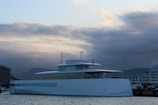 A sleek, modern yacht is docked at a marina, surrounded by calm waters. The yacht has a minimalist design with large windows along its side. In the background, there are partially cloudy skies with hints of a setting sun, casting a serene glow over the scene. Buildings are visible on the left and right, suggesting an urban waterfront location.