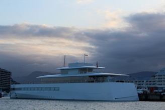 A sleek, modern yacht is docked at a marina, surrounded by calm waters. The yacht has a minimalist design with large windows along its side. In the background, there are partially cloudy skies with hints of a setting sun, casting a serene glow over the scene. Buildings are visible on the left and right, suggesting an urban waterfront location.