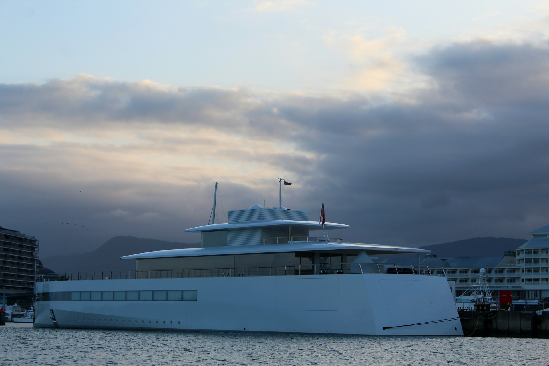 A sleek, modern yacht is docked at a marina, surrounded by calm waters. The yacht has a minimalist design with large windows along its side. In the background, there are partially cloudy skies with hints of a setting sun, casting a serene glow over the scene. Buildings are visible on the left and right, suggesting an urban waterfront location.