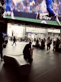 A busy public space with a large digital screen displaying a football (soccer) match. The area is filled with people walking and sitting, some carrying bags. One person is seated on a modern, curved bench, reading a book. In the background, there is the entrance to Tottenham Court Road Station, with multiple people gathered around.