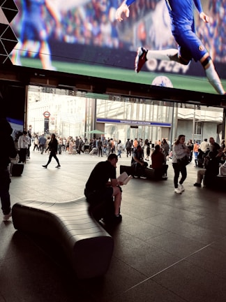 A busy public space with a large digital screen displaying a football (soccer) match. The area is filled with people walking and sitting, some carrying bags. One person is seated on a modern, curved bench, reading a book. In the background, there is the entrance to Tottenham Court Road Station, with multiple people gathered around.