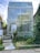 A modern, rectangular building constructed with glass blocks is surrounded by lush greenery and overgrown plants. A small ramp with handrails leads up to the building's entrance, adding an element of accessibility. The sky is partially cloudy, creating a soft background.