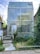 A modern, rectangular building constructed with glass blocks is surrounded by lush greenery and overgrown plants. A small ramp with handrails leads up to the building's entrance, adding an element of accessibility. The sky is partially cloudy, creating a soft background.