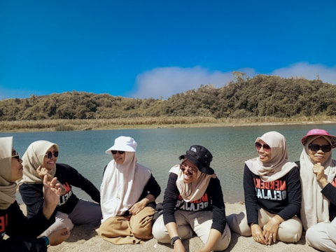 A group of women wearing matching outfits sit on a sandy beach near a body of water, with green hills and a clear blue sky in the background. Some are wearing hats and sunglasses, and they appear to be engaged in conversation.