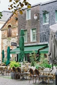 A charming outdoor café with an arrangement of woven chairs and tables, situated on a cobblestone path. The café exterior features rustic brick and wooden shingle walls, alongside several green umbrellas and potted plants adding to the verdant atmosphere. The ambiance suggests a European setting with ivy-covered walls and classic lantern-style lighting fixtures.