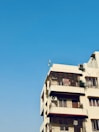 A multi-story residential building with a beige facade, featuring several balconies with ornate metal railings. An air conditioning unit is attached to one of the balconies, and a small green plant is visible on another. A flag is mounted on the roof, and the background is a clear blue sky.