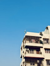 A multi-story residential building with a beige facade, featuring several balconies with ornate metal railings. An air conditioning unit is attached to one of the balconies, and a small green plant is visible on another. A flag is mounted on the roof, and the background is a clear blue sky.