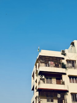A multi-story residential building with a beige facade, featuring several balconies with ornate metal railings. An air conditioning unit is attached to one of the balconies, and a small green plant is visible on another. A flag is mounted on the roof, and the background is a clear blue sky.
