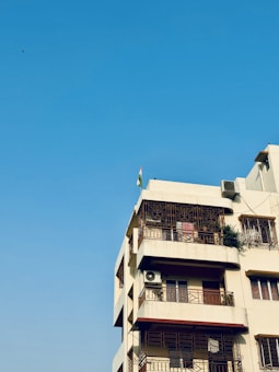 A multi-story residential building with a beige facade, featuring several balconies with ornate metal railings. An air conditioning unit is attached to one of the balconies, and a small green plant is visible on another. A flag is mounted on the roof, and the background is a clear blue sky.