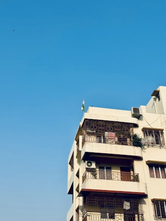 A multi-story residential building with a beige facade, featuring several balconies with ornate metal railings. An air conditioning unit is attached to one of the balconies, and a small green plant is visible on another. A flag is mounted on the roof, and the background is a clear blue sky.