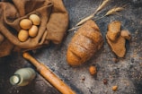 A rustic table setting with a loaf of seeded brown bread, several slices of the bread, a bunch of wheat stalks, a fabric sack containing brown eggs, a vintage milk bottle, and a wooden rolling pin. The background features a wooden surface sprinkled with flour and scattered nuts and seeds.