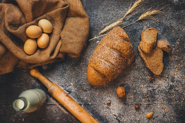 A rustic table setting with a loaf of seeded brown bread, several slices of the bread, a bunch of wheat stalks, a fabric sack containing brown eggs, a vintage milk bottle, and a wooden rolling pin. The background features a wooden surface sprinkled with flour and scattered nuts and seeds.