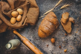 A rustic table setting with a loaf of seeded brown bread, several slices of the bread, a bunch of wheat stalks, a fabric sack containing brown eggs, a vintage milk bottle, and a wooden rolling pin. The background features a wooden surface sprinkled with flour and scattered nuts and seeds.