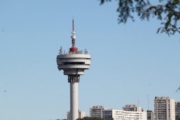 A tall telecommunications tower stands prominently against a clear blue sky, featuring a cylindrical structure with multiple tiers and various antennas on top. In the background, several high-rise residential buildings form a cityscape beneath the tower. At the edge of the frame, tree branches add a natural element to the scene.