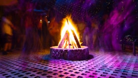 A vibrant campfire surrounded by blurred silhouettes of people, creating a dynamic and festive atmosphere. The ground is covered with a patterned surface, and colorful lights illuminate the scene with hues of purple and yellow.